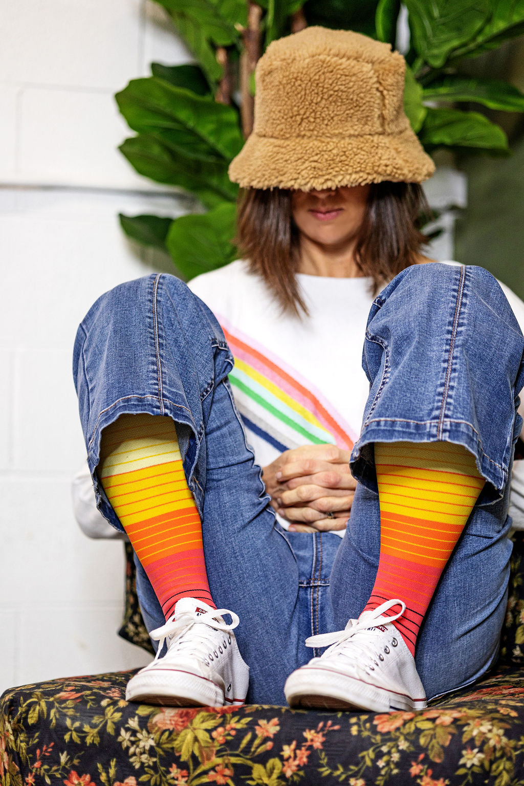 Woman on a couch wearing Lunatik Retro Renegade+ compression socks (rainbow stripes) with white canvas shoes..